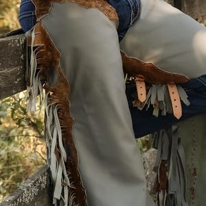 May include: Gray leather chaps with brown fringe and accents. The chaps have a brown, textured upper section with a buckle closure. The chaps are worn over blue jeans and are suitable for horseback riding or Western-themed events.