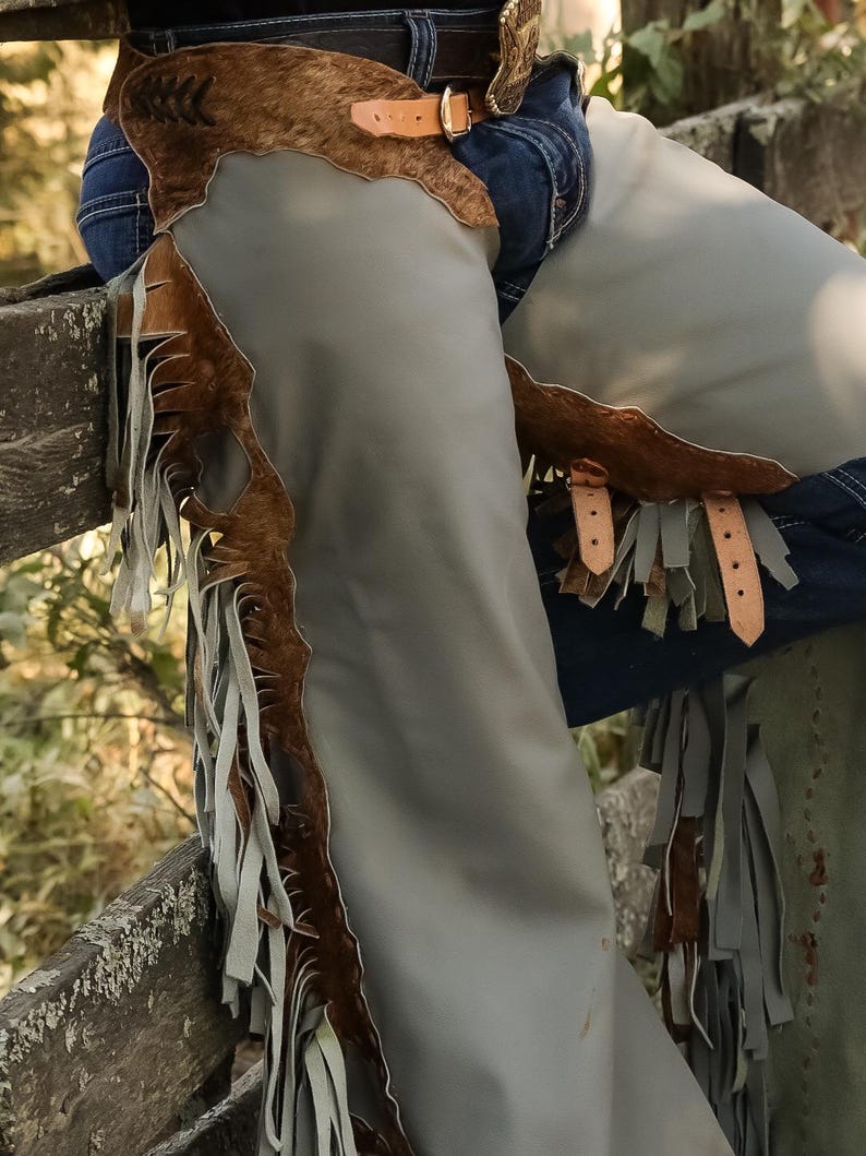 May include: Gray leather chaps with brown fringe and accents. The chaps have a brown, textured upper section with a buckle closure. The chaps are worn over blue jeans and are suitable for horseback riding or Western-themed events.