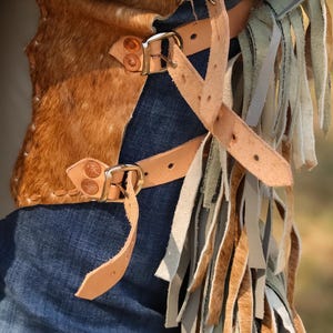 May include: Brown and tan leather chaps with a cowhide panel and fringe detailing. The chaps feature adjustable leather straps with metal buckles and are worn over blue denim jeans. The fringe includes shades of tan, gray, and green.