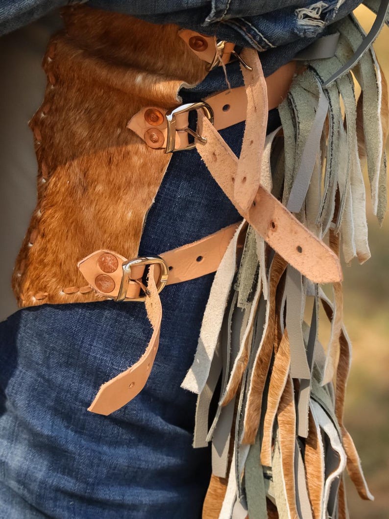 May include: Brown and tan leather chaps with a cowhide panel and fringe detailing. The chaps feature adjustable leather straps with metal buckles and are worn over blue denim jeans. The fringe includes shades of tan, gray, and green.
