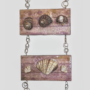 Puede incluir: Un colgante de pared hecho de cuatro placas de madera con conchas marinas unidas a cada una. Las placas están conectadas por una cadena plateada. Las conchas son de varios tonos de marrón y blanco.