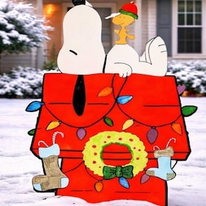 May include: A red wooden Snoopy doghouse Christmas decoration featuring Snoopy and Woodstock. The doghouse is adorned with colourful lights, a green wreath, and stockings. The scene is set in a snowy outdoor environment.