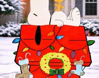 May include: A red wooden Snoopy doghouse Christmas decoration featuring Snoopy and Woodstock. The doghouse is adorned with colourful lights, a green wreath, and stockings. The scene is set in a snowy outdoor environment.