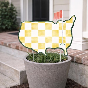 Op de afbeelding: Een decoratief tuinschild in de vorm van de Verenigde Staten, met een geel en wit geblokt patroon. Het heeft een groene rand en een kleine rode vlag. Het bord staat in een grijze betonnen plantenbak gevuld met groen gras.