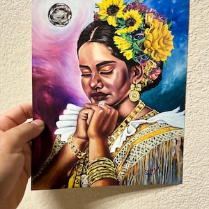 Puede incluir: Una pintura colorida de una mujer con los ojos cerrados, adornada con un tocado floral y joyas de oro. La obra de arte presenta una paleta de colores vibrantes con un elemento celestial en el fondo. Las manos de la mujer están juntas en oración.