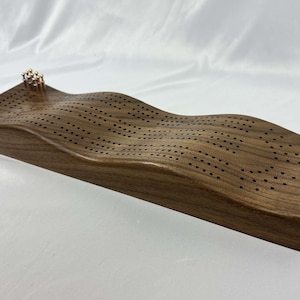 Peut inclure: Un plateau de jeu de cribbage en bois avec une surface ondulée et des trous pour les pions. Le plateau est en bois foncé et a une finition naturelle.