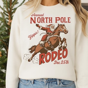 May include: Cream-colored sweatshirt with a festive graphic. The design features Santa Claus riding a horse in a rodeo, with the text "Annual North Pole Rodeo Dec. 25th" in red. The word "Yippie!" is also included.