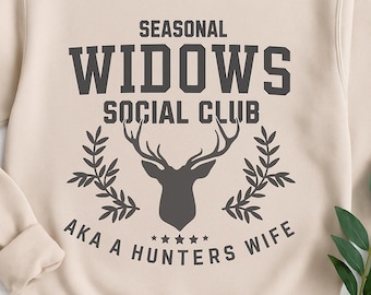 Pode incluir: Camisola bege de gola redonda com o texto "SEASONAL WIDOWS SOCIAL CLUB AKA A HUNTERS WIFE" em cinza escuro. O design inclui uma silhueta de cabeça de veado e ramos de louro.