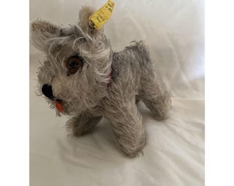 Può includere: Un giocattolo di peluche vintage a forma di cane grigio e peloso con un collare rosso e un'etichetta gialla che recita "Made in W. Germany" e "Schuco Spielwaren Germany".