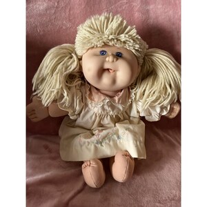 May include: A vintage Cabbage Patch Kid doll with blonde yarn hair, blue eyes, and a pink and white dress. The doll is sitting on a pink surface.