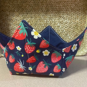 May include: A fabric bowl cosy featuring a dark blue background with a repeating pattern of red strawberries, green leaves, and small white flowers with yellow centres. The bowl cosy has a square base and four pointed corners with visible stitching.
