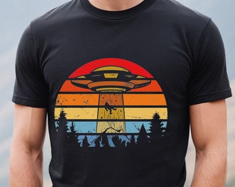 Könnte beinhalten: Ein schwarzes T-Shirt mit einer Retro-Grafik eines UFOs, das über eine Bergkette mit einem Sonnenuntergang im Hintergrund fliegt.