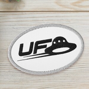 May include: White oval patch with black embroidered text "UFO" and a black UFO graphic. Two potted green succulent plants are on a white wooden surface.