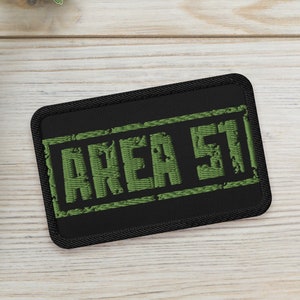 May include: A black patch with white stitching and the text "AREA 51" in green. The patch is on a light wood surface with two potted succulents.