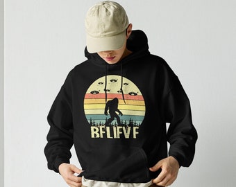 May include: A black hooded sweatshirt with a vintage-style graphic of a Bigfoot walking with UFOs above him. The graphic has a rainbow color scheme and the text "BELIEVE" is printed below the graphic. The model is wearing khaki cargo pants and white sneakers.
