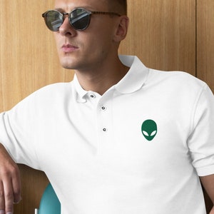 May include: White polo shirt with a green alien head graphic on the left chest. The person is wearing sunglasses and sitting on a teal chair.
