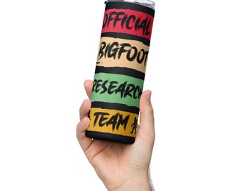 May include: A black stainless steel tumbler with a colorful design. The tumbler has four horizontal stripes in red, beige, green, and yellow. The text "OFFICIAL BIGFOOT RESEARCH TEAM" is printed in black on the stripes.