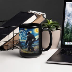 May include: Black ceramic mug with a white handle. The mug features a colorful image of a Bigfoot walking through a starry night landscape. The image is reminiscent of Vincent van Gogh's Starry Night.