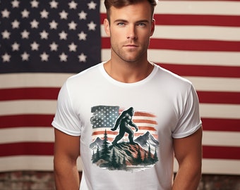 Puede incluir: Una camiseta blanca con un gráfico de Bigfoot caminando por una montaña con una bandera estadounidense en el fondo.