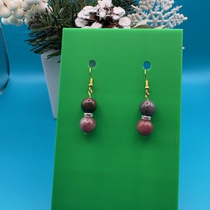 May include: A pair of dangle earrings with gold-colored hooks. Each earring features two round, polished beads: a dark purple bead at the top and a reddish-brown bead below, separated by a silver-colored spacer. The earrings are displayed on a green background.
