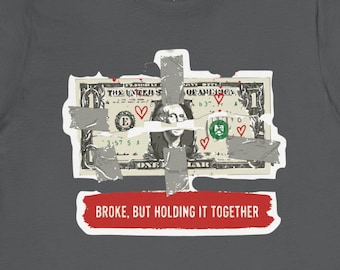 May include: Dark grey t-shirt featuring a graphic of a dollar bill design with tape and hearts. The text "BROKE, BUT HOLDING IT TOGETHER" is printed on a red banner below the dollar bill graphic.