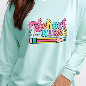 May include: A light teal long-sleeve shirt with the words "School Nurse" in colorful, bubble letters. Below the text is a pencil and a heartbeat line. The shirt is made of a soft material.
