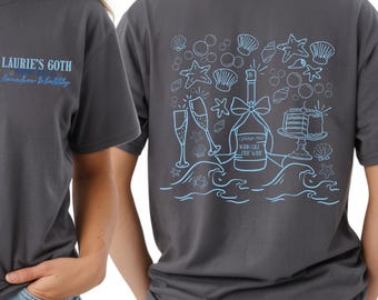 May include: Charcoal gray t-shirt with the text "Laurie's 60th, Sunshine & Bubbly" on the front. The back features a light blue illustration of champagne, cake, and seashells. A casual, short-sleeved tee.