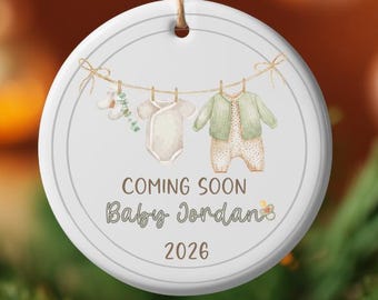 May include: A white ceramic ornament with a watercolor design. The ornament features baby clothes hanging on a clothesline, with the text "COMING SOON Baby Jordan 2026". The ornament is hanging from a brown string.