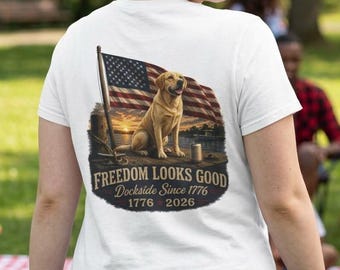 May include: White t-shirt with a graphic featuring a yellow Labrador Retriever, an American flag, and the text "Freedom Looks Good Dockside Since 1776 1776 2026". The shirt is worn outdoors.