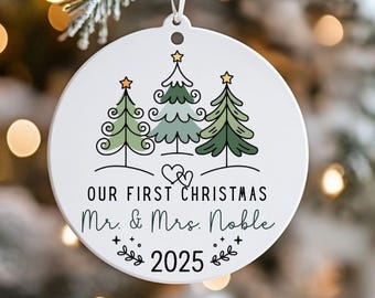 May include: White round Christmas ornament with three green Christmas trees and gold stars. The text "OUR FIRST CHRISTMAS Mr. & Mrs. Noble 2025" is printed on the ornament. The ornament hangs from a white ribbon and is displayed on a Christmas tree.