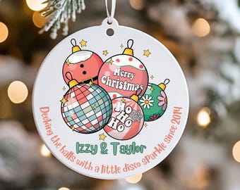 May include: A white Christmas ornament with a festive design. The ornament features colorful disco ball-style ornaments with the text "Merry Christmas" and "Ho Ho Ho." The ornament also includes the text "Izzy & Taylor" and "Decking the halls with a little disco sparkle since 2014."