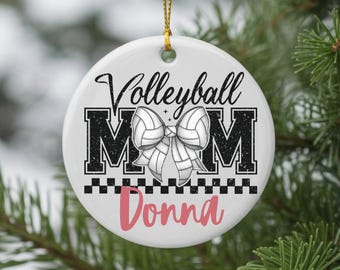 May include: White round ceramic ornament with the words "Volleyball MOM" in black and a volleyball bow design. The name "Donna" is in pink script below. A gold string is attached for hanging.