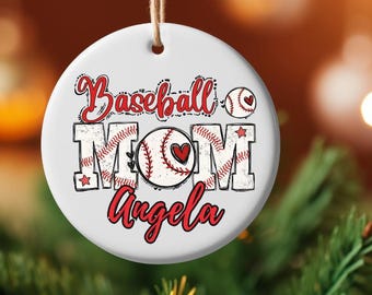 May include: A white, round Christmas ornament with the words "Baseball MOM Angela" in red and black lettering. The word "MOM" is in a baseball-themed font. A baseball graphic with a heart is in the center. The ornament hangs from a brown string.