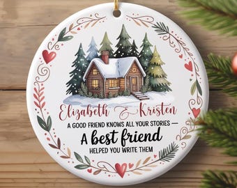 May include: A round, white Christmas ornament with a watercolor illustration of a cozy cabin in a snowy forest. The ornament has text that reads "Elizabeth & Kristen" and "A best friend helped you write them."