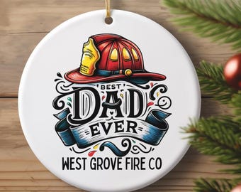 May include: A white, round Christmas ornament with a firefighter's helmet design. The ornament reads "Best Dad Ever" with a blue ribbon banner and the text "West Grove Fire Co."