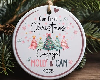 May include: A white ceramic Christmas ornament with the text "Our First Christmas Engaged" and the names "Molly & Cam" with the year 2025. The ornament features a design of Christmas trees in pink, green, and blue, with a brown string for hanging.