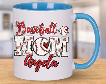 May include: White ceramic mug with a blue interior and handle. The mug features the words "Baseball MOM Angela" in red script and block letters, with baseball graphics. A heart is in the center of the "O" in MOM. The mug is on a wooden surface.