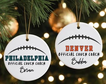 May include: Two white ceramic Christmas ornaments hanging from a Christmas tree. Each ornament has a football design and text. One says "PHILADELPHIA Official Couch Coach Brian" and the other says "DENVER Official Couch Coach Bubba".