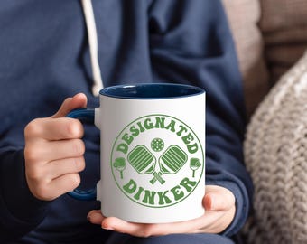 May include: White ceramic mug with a navy blue interior and handle. The mug features a green circular design with the words "DESIGNATED DINKER" around a pickleball and crossed paddles.