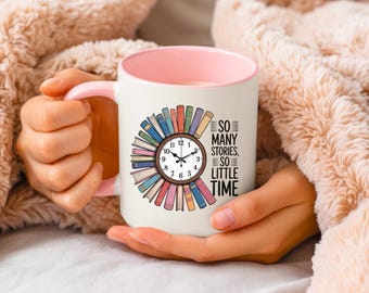 May include: White ceramic mug with a pink interior and handle. The mug features a clock design surrounded by colorful books. Text on the mug reads "SO MANY STORIES, SO LITTLE TIME". The mug is held by a person.
