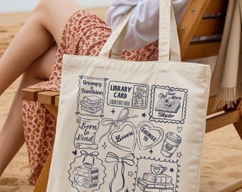 May include: Cream-colored tote bag with a navy blue book-themed design. The bag features illustrations of books, hearts, and coffee cups, along with the text "Grumpy x Sunshine," "Library Card," "Born to Read," "Lovers," "Enemies to," "Just One More Chapter," "Amanda," and "Booked & Busy."