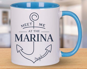 May include: White ceramic mug with a blue interior and handle. The mug features the text "MEET ME AT THE MARINA" with an anchor graphic in navy blue. The name "Angela" is written below the anchor.