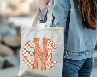 May include: Cream-colored tote bag with a large, colorful monogram design. The monogram features the letters "GNI" in a retro style, with floral, checkered, and patterned elements in shades of orange, brown, and pink. The bag has long straps.