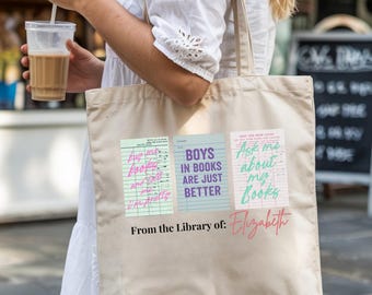 May include: A beige canvas tote bag featuring three library card-themed designs with text. The text includes "Ask me about my books", "Boys in books are just better", and "From the Library of: Elizabeth". The bag is carried by a person.