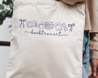 May include: Cream-colored tote bag with a blue line drawing of books, a coffee cup, glasses, and bows. The word "booktrovert" is written in cursive below the illustrations. The bag is held by a person.