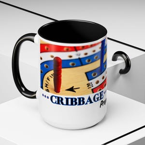 May include: White ceramic mug with a black handle and rim. The mug features a colorful image of a cribbage board game with a red peg and the text "...CRIBBAGE...Pro"