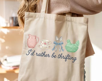 May include: Beige tote bag with a vintage-style illustration. The design includes a pink pitcher, a teacup with a floral pattern, silverware tied with a blue bow, and a green hen. The text "I'd rather be thrifting" is written in a script font.