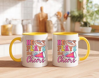 May include: Two white coffee mugs with yellow interiors and handles. Each mug features the text "HE PLAYS she Cheers" with a football and a cheerleader megaphone. The mugs are on a wooden surface.