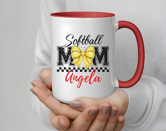 May include: White ceramic mug with a red interior and handle. The mug features the text "Softball Mom" with a yellow bow graphic and the name "Angela" in red. A black and white checkered pattern is at the bottom of the design.