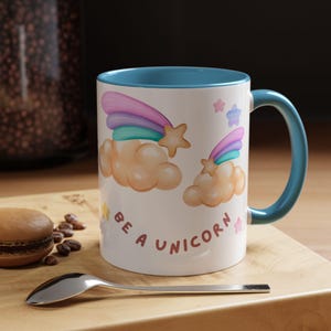 May include: White ceramic mug with a blue rim. The mug features a rainbow and clouds with stars. The text "Be a Unicorn" is printed on the mug.